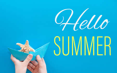 Hello summer. Summer card with a paper boat and a starfish on a blue background in the hands of a child. Travel to the sea and vacation in summerの写真素材