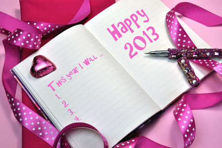 2013 Happy New Year message in pink diary with New Year Resolutions list on pink background with pink heart chocolate, polka dot ribbon and pink bling writing pen.の写真素材
