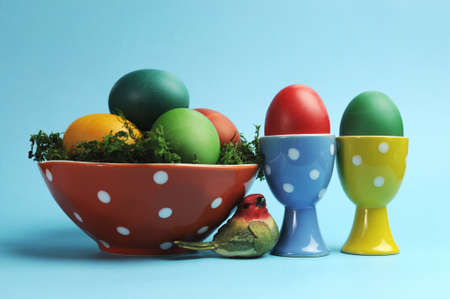 Bright and cheerful Happy Easter still life with rainbow color eggs in orange polka dot bowl and egg cups against a blue background の写真素材