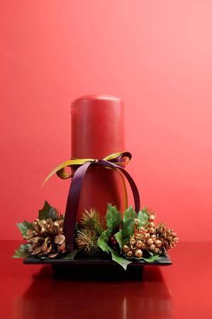 Red candle with holly and pine cone Christmas table centrepiece ornament against a festive holiday red background の写真素材