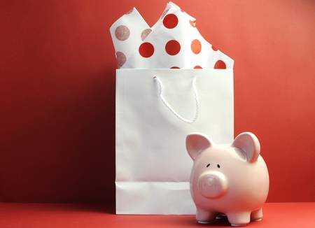 Savings concept with white shopping bag, red polka dot tissue paper, and cute pink piggy bank against a red background の写真素材