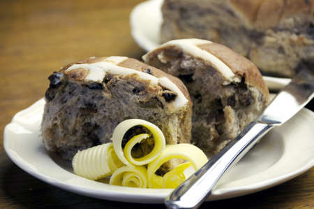 Close-up of traditional Easter treat, Hot Cross Buns, on awhite plate with knife and butter curls, on a wood table background.の写真素材