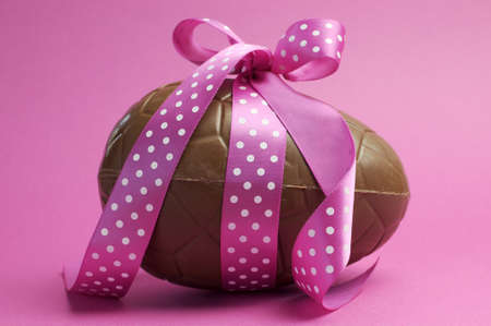 Large Happy Easter chocolate Easter egg with pink polka dot ribbon tied in a bow against a pretty feminine pink background の写真素材