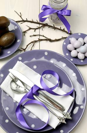 Purple theme Easter dinner, breakfast or brunch table setting with chocolate bunny rabbit, and sugar candy coated birds nest eggs on polka dot plates on white shabby chic natural wood table  Aerial vertical orientation の写真素材