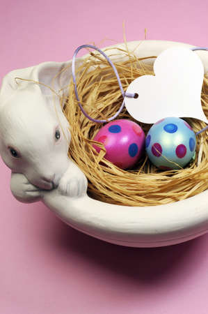 Pink and blue Easter eggs in white bunny bowl with heart gift tag on pink background  Vertical 