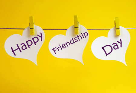 Celebrate International Friendship Day with Happy Friendship Day message across three white heart tags hanging from pegs on a line against a yellow background の写真素材