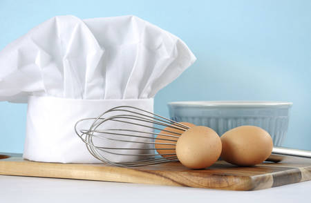 Modern kitchen cooking kitchenware and chef's hat with mixing bowl, whisk, chopping boards and eggs on a pale aqua blue and white background. Close up with copy space.の写真素材