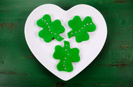 Happy St Patricks Day shamrock shape green fondant cookies on white heart shape plate on vintage style green wood table.の写真素材