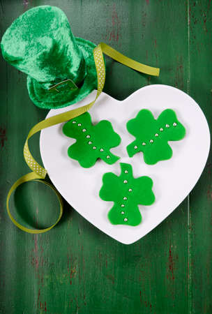 Happy St Patricks Day shamrock shape green fondant cookies on white heart shape plate on vintage style green wood table.の写真素材