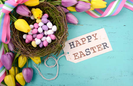 Happy Easter background with painted Easter eggs in birds nest, and yellow and purple silk tulips and ribbon on vintage style rustic distressed aqua blue wood table, with greeting tag.の写真素材