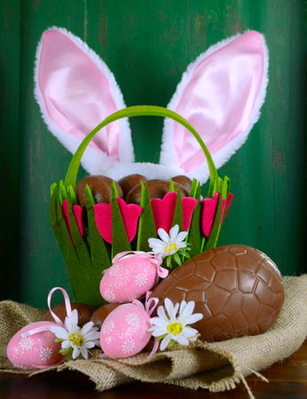Happy Easter basket with bunny ears of chocolate Easter eggs on burlap canvas against a green wood...