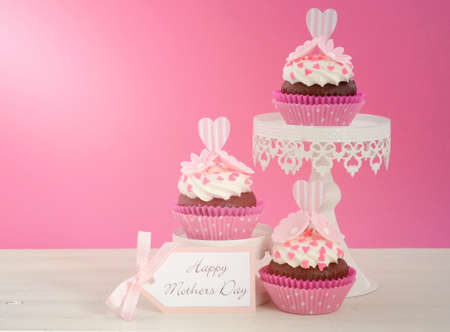 Happy Mothers Day pink and white cupcakes on retro style cake stands on vintage white wood table,...