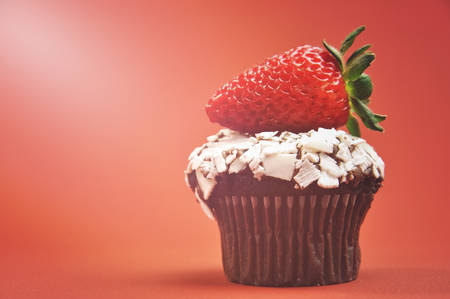 Close-up of a delicious red strawberry on a mini chocolate muffin cupcake against a red orange background, with copy space and applied retro style filters and added lens flare light beam.の写真素材