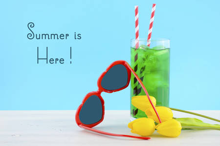 Summer Is Here concept with red heart shape sunglasses and yellow flowers and tall cool drink on white wood table and sky blue background.の写真素材