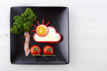 Bright happy breakfast with sunny egg, bacon tree with parsley leaves and cherry tomato flowers on black square plate on white wood table.の写真素材