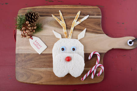 Christmas fun for the children with reindeer face cut out of bread sandwich with decorations on chopping board on a rustic red wood table.の写真素材