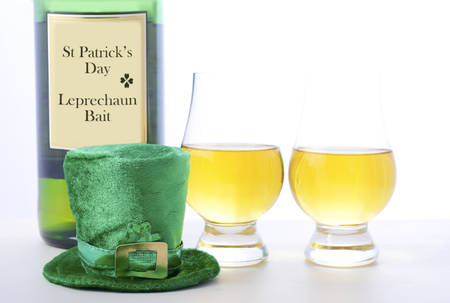 St Patricks Day Irish Whisky bottle with glasses and leprechaun hat on white wood table.