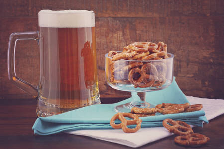 Glass of beer in stein with mini pretzels on pale blue napkin on wood background, with applied retro style filters.の写真素材