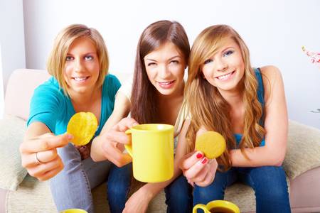 Three happy friends beautiful girls sitting on sofa in home with cups, chips and cookieの写真素材