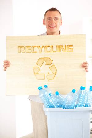 Happy man holding wooden board with word recycling and bin full of plastic blue bottlesの写真素材