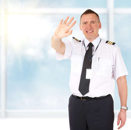Cheerful airline pilot wearing uniform with epaulettes sending greets with hand, standing at the airports big windowの写真素材