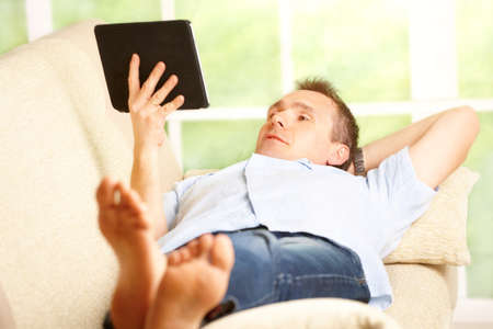 Man relaxing with tablet, laying on sofa in home with big window in backgroundの写真素材