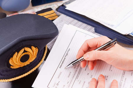 Close up of an airplane pilot hand filling in an flight plan with equipment including hat,...