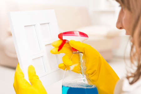 Beautiful young woman cleaning frame in her home wearing yellow gloves and using cleaning sprayの写真素材