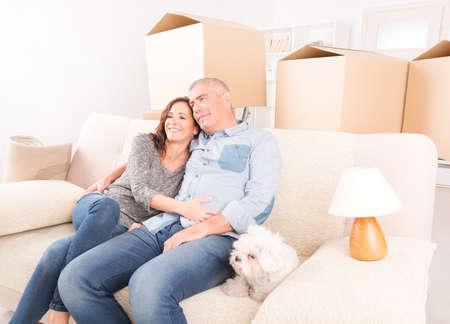 Happy mature couple celebrating their new home sitting together on the sofa with their little dog ...