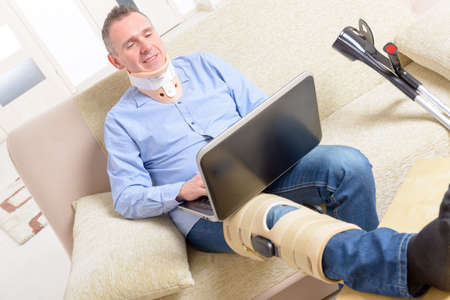 Man with leg in neck brace, knee cages and crutches for stabilization and support resting on a sofa with laptop.の写真素材