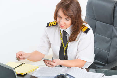 Beautiful woman pilot wearing uniform with epaulettes using mobile phone and laptop at preflight...
