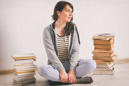 Woman listening an audiobook on smartphone sitting between piles of paper books on the floorの写真素材