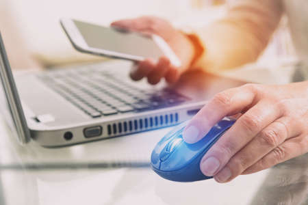 Hand on computer mouse and smartphone in the other. notebook in background in deskの写真素材