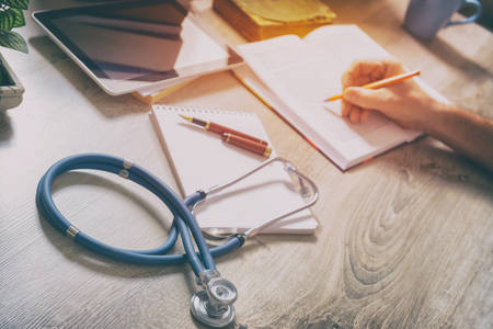 Young doctor is learning and preparing for the exam, she is using books and tablet to search for...