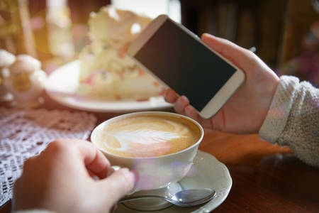 Authentic image of woman using smart phone taken in the cafe with cappuccino and meringue cake in the backgroundの写真素材