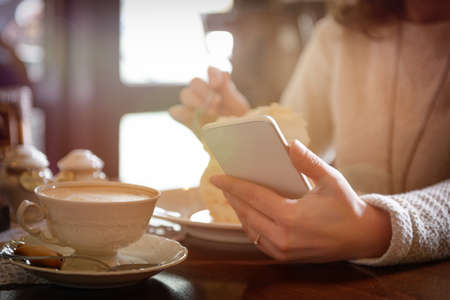 Authentic image of woman using smart phone taken in the cafe with cappuccino and meringue cake in...