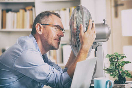 Man suffers from heat while working in the office and tries to cool off by the fanの写真素材
