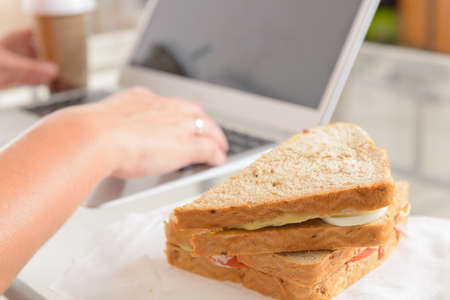 Woman eating a breakfast sandwich and drinking coffee while working with a laptopの写真素材