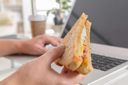 Woman eating a breakfast sandwich and drinking coffee while working with a laptopの写真素材