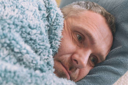 Mature man suffering from depression lies sadly covered with a blanket on his bedの写真素材