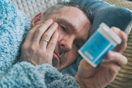 Mature man suffering from depression lies sadly covered with a blanket on his bed and holds a bottle...