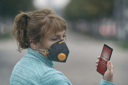 Woman wearing a real anti-pollution, anti-smog and viruses face mask and checking current air pollution with smart phone appの写真素材