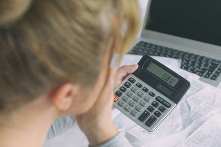 Woman uses a calculator to sum up expenses and holds her head worrying about the amount of money...