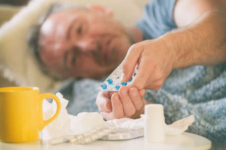 Man suffering from coronavirus, flu or cold lies sadly covered with a blanket on his bed. Drugs,...