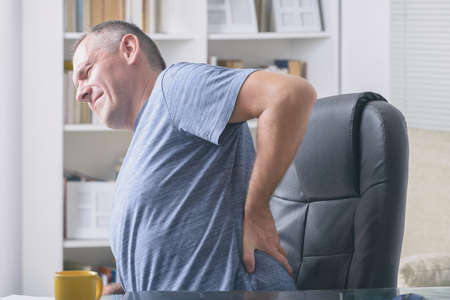 Man in home office suffering from low back pain sitting at a desk with notebook, papers and other...