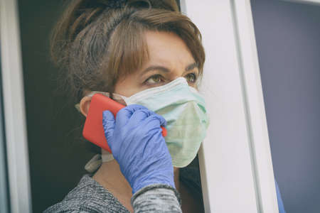 Woman using smartphone wearing a protective face mask and looking out the window of her home because of an epidemic of corona virus covid-19. Home quarantine and self isolation conceptの写真素材