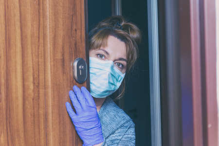 Woman wearing a protective face mask and gloves opening doors of her home because of an epidemic of...