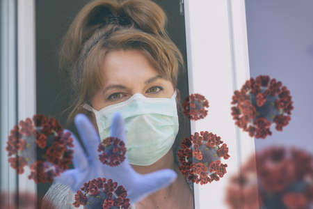 Woman wearing a protective face mask and looking out the window of her home because of an epidemic of corona virus covid-19. Home quarantine and self isolation conceptの写真素材