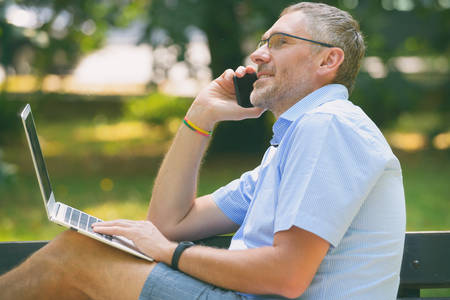 Businessman holding his laptop using his smartphone, sitting in park
