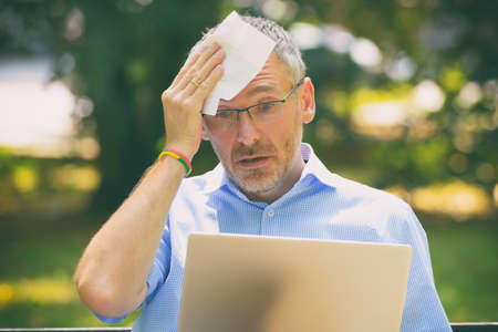 Man suffers from heat while working with laptop in the park and wiping his forehead with tissueの写真素材